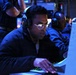 The image shows personnel working at computer workstations in a dimly lit, blue-toned operations or command center environment. The foreground subject is wearing headphones with a boom microphone and glasses, actively typing on a keyboard with illuminated green keys. The setting appears to be a military or naval combat information center (CIC) or similar tactical operations room.