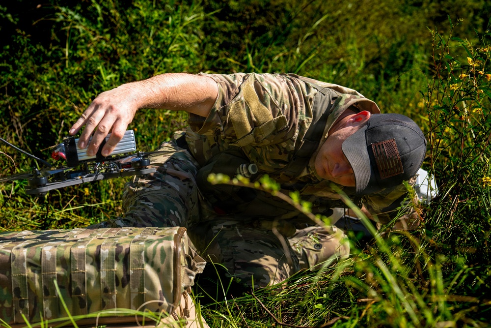 A person wearing military-style multicam camouflage clothing and a baseball cap with a flag patch is crouched low in a grassy, vegetated area. They are handling a small drone or FPV quadcopter, appearing to prepare it for launch or retrieve it. A multicam-patterned bag or case is visible on the ground nearby.