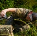 A person wearing military-style multicam camouflage clothing and a baseball cap with a flag patch is crouched low in a grassy, vegetated area. They are handling a small drone or FPV quadcopter, appearing to prepare it for launch or retrieve it. A multicam-patterned bag or case is visible on the ground nearby.