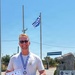 A smiling man with blonde hair and sunglasses stands outdoors holding a diploma/certificate and wearing a medal on a blue and white ribbon. He is wearing a white t-shirt with 'USS TUSCON SSN 770' printed on it and has a teal/green ribbon draped over his arm. A Greek flag flies on a flagpole behind him, and a stone monument with Greek text is visible to his right.