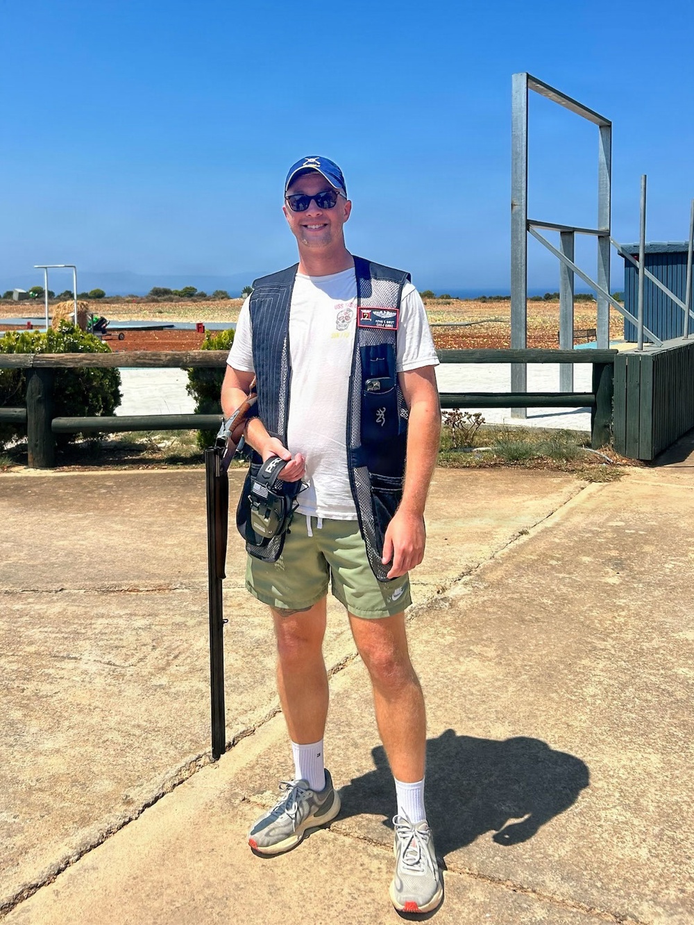 A man stands at what appears to be a clay pigeon or trap shooting range on a sunny day. He is equipped with a shotgun, a shooting vest with a Browning logo, and an ammunition pouch. The background shows an open shooting range with flat terrain and a blue sky.