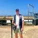 A man stands at what appears to be a clay pigeon or trap shooting range on a sunny day. He is equipped with a shotgun, a shooting vest with a Browning logo, and an ammunition pouch. The background shows an open shooting range with flat terrain and a blue sky.