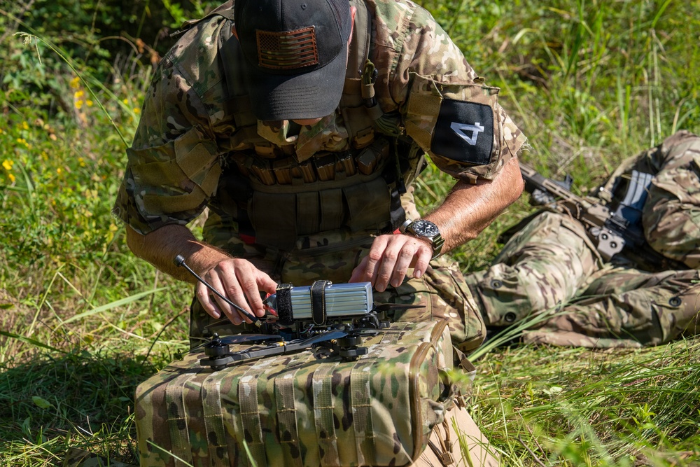 A military personnel wearing multicam camouflage uniform and a baseball cap with a US flag patch is kneeling in a grassy outdoor area, assembling or configuring what appears to be a small quadcopter drone on top of a camouflage bag. A second individual in similar military attire is visible in the background lying in a prone position with a weapon. The setting appears to be a field training exercise.