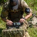 A military personnel wearing multicam camouflage uniform and a baseball cap with a US flag patch is kneeling in a grassy outdoor area, assembling or configuring what appears to be a small quadcopter drone on top of a camouflage bag. A second individual in similar military attire is visible in the background lying in a prone position with a weapon. The setting appears to be a field training exercise.