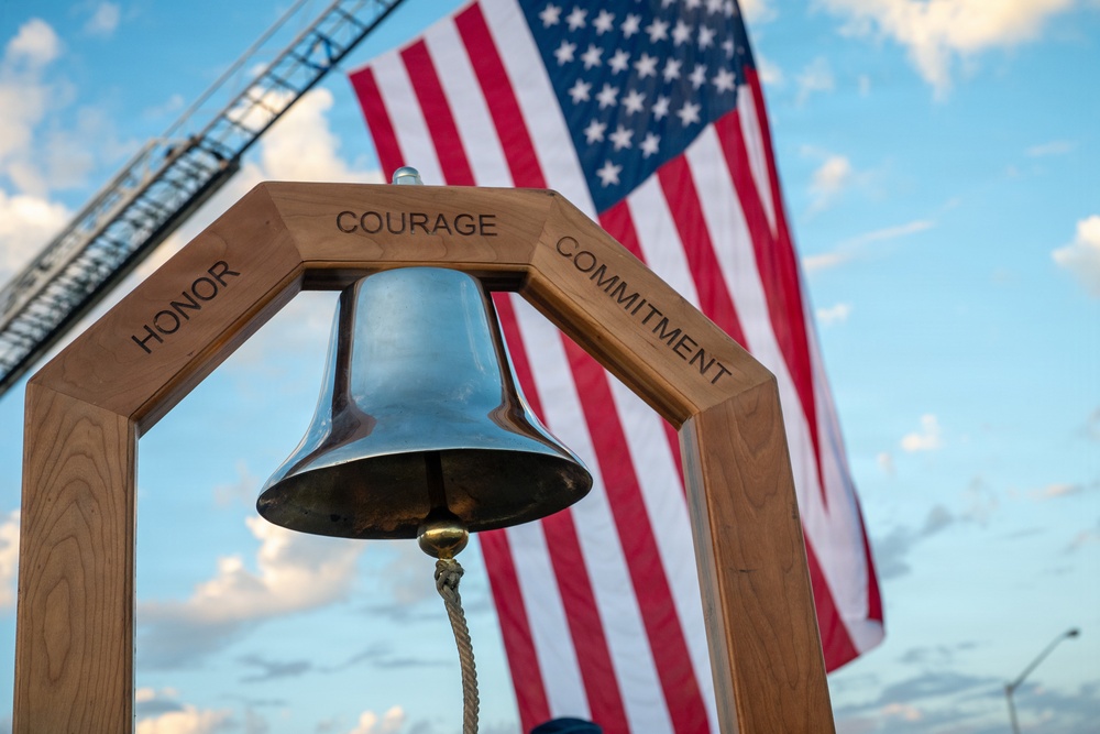 A polished metal bell hangs from a wooden A-frame stand engraved with the words 'Honor,' 'Courage,' and 'Commitment.' The bell is displayed against a large American flag and a partly cloudy sky, with what appears to be a crane or ladder structure visible in the background. This appears to be a ceremonial or memorial setting, likely associated with military, firefighting, or first responder commemoration.