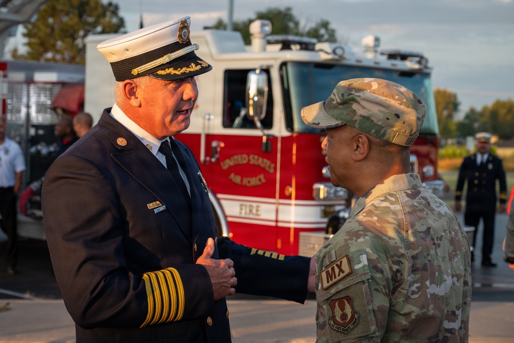 A senior fire official in dress uniform is engaged in conversation with a U.S. Air Force military member in OCP (Operational Camouflage Pattern) uniform. The interaction takes place in front of a United States Air Force Fire Department fire truck, suggesting a formal or ceremonial military event. Additional personnel in dress uniforms are visible in the background.