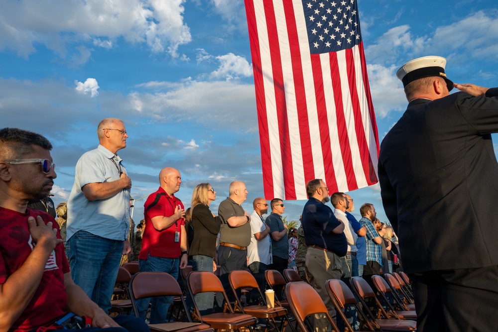 An outdoor patriotic ceremony is taking place with a large American flag prominently displayed. Attendees are standing with hands over their hearts, suggesting the playing of the National Anthem or Pledge of Allegiance. A uniformed military or naval officer in a dark dress uniform and white cap is saluting on the right side of the frame.