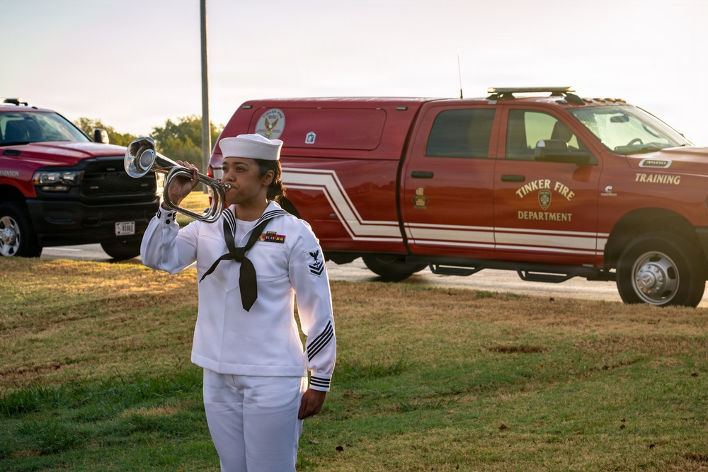 A U.S. Navy sailor in dress white uniform plays a bugle during what appears to be a military ceremony or memorial event. The setting is outdoors on a grassy area, with a Tinker Fire Department training truck prominently visible in the background. The lighting suggests either early morning or late afternoon, consistent with a formal military observance such as Taps or Reveille.
