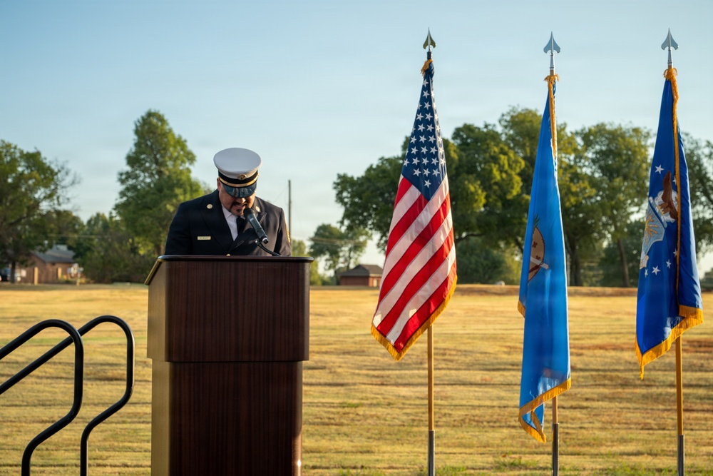 A uniformed official, appearing to be a fire department officer based on his dress uniform and white cap, speaks at a wooden podium during an outdoor ceremony. Three flags are displayed behind him: the United States flag, what appears to be the Oklahoma state flag, and a U.S. Air Force flag. The ceremony takes place on a grassy field during golden hour lighting, suggesting an early morning or late afternoon event.