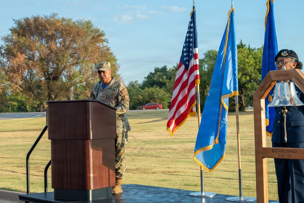 A military ceremony is taking place outdoors, featuring a uniformed service member speaking at a wooden podium. Three flags are displayed — the U.S. flag, what appears to be the Oklahoma state flag, and a U.S. Air Force flag — alongside a ceremonial bell on a stand inscribed with 'HONOR, COURAGE, COMMITMENT' and 'ESTABLISHED 29 MAY 1992'. A second uniformed individual stands near the bell on the right side of the stage.