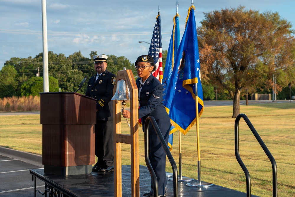 A formal military or civic memorial ceremony is taking place on an outdoor stage. A female Air Force airman in dress blues is ringing a ceremonial bell mounted on a wooden stand, while a fire department official in dress uniform stands at a podium behind her. Three flags — the U.S. flag and two blue military/organizational flags — are displayed on the stage.