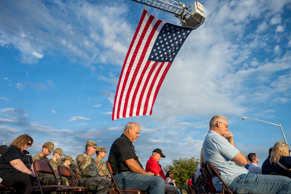 An outdoor patriotic ceremony or memorial event featuring a large American flag suspended from what appears to be a fire truck ladder aerial arm. A crowd of attendees, including uniformed military personnel in camouflage uniforms, are seated in folding chairs with heads bowed, suggesting a moment of prayer or reflection. The scene is set against a partly cloudy blue sky during what appears to be late afternoon or early evening light.