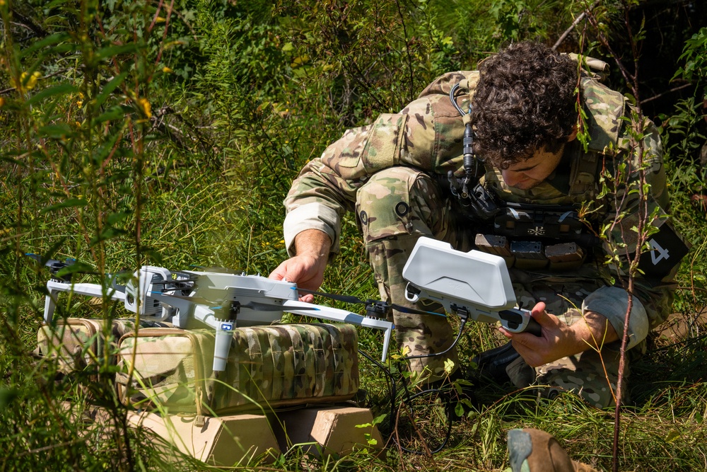 A soldier in multicam/OCP camouflage uniform is crouched in a vegetated outdoor area, preparing or assembling a small commercial-style quadcopter drone. The drone appears to be white and is resting on what looks like military-patterned equipment bags. The setting suggests a field training exercise or operational preparation in a wooded or brushy environment.