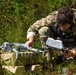 A soldier in multicam/OCP camouflage uniform is crouched in a vegetated outdoor area, preparing or assembling a small commercial-style quadcopter drone. The drone appears to be white and is resting on what looks like military-patterned equipment bags. The setting suggests a field training exercise or operational preparation in a wooded or brushy environment.