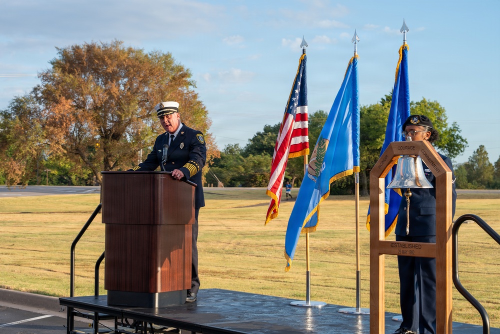 An outdoor memorial or ceremonial event featuring a fire department official in dress uniform speaking at a wooden podium on a raised stage. Three flags are displayed — the United States flag, what appears to be the Oklahoma state flag, and a blue military or organizational flag. A second uniformed individual stands beside a ceremonial bell on a dedicated stand to the right.