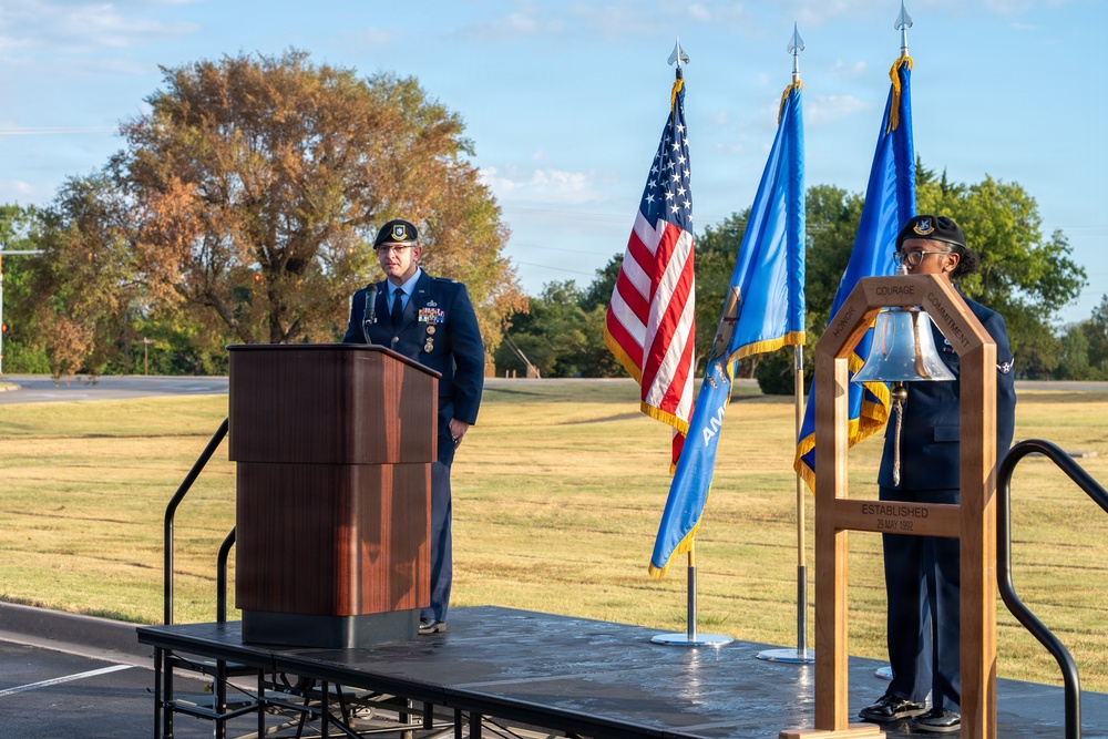 A formal military ceremony is taking place on an outdoor stage, featuring two uniformed Air Force personnel. One officer stands at a wooden podium delivering remarks while another stands beside a ceremonial bell. Three flags — the U.S. flag, what appears to be an Oklahoma state flag, and a U.S. Air Force flag — are displayed prominently on the stage.