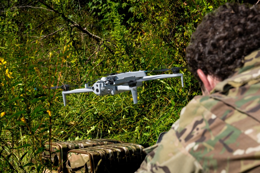 A person wearing military camouflage (MultiCam/OCP pattern) is operating a quadcopter drone in an outdoor wooded environment. The drone, appearing to be a DJI Mavic 3 Enterprise or similar model, is hovering at low altitude in front of dense green vegetation. A camouflage-patterned equipment case or bag is visible in the foreground.
