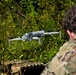 A person wearing military camouflage (MultiCam/OCP pattern) is operating a quadcopter drone in an outdoor wooded environment. The drone, appearing to be a DJI Mavic 3 Enterprise or similar model, is hovering at low altitude in front of dense green vegetation. A camouflage-patterned equipment case or bag is visible in the foreground.