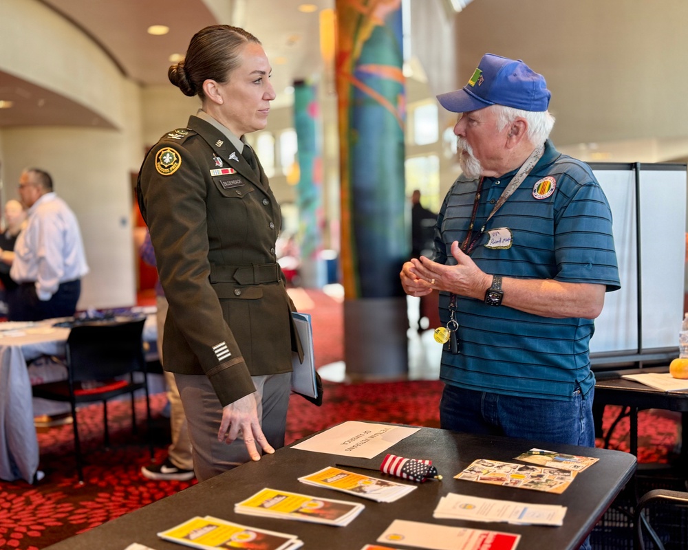 A U.S. Army soldier in dress green uniform is engaged in conversation with an older civilian man wearing a blue cap and striped polo shirt at what appears to be a veterans outreach or recruitment event. They are standing at a table covered with brochures, pamphlets, and informational materials. The setting is an indoor venue with decorative columns and additional attendees visible in the background.