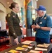 A U.S. Army soldier in dress green uniform is engaged in conversation with an older civilian man wearing a blue cap and striped polo shirt at what appears to be a veterans outreach or recruitment event. They are standing at a table covered with brochures, pamphlets, and informational materials. The setting is an indoor venue with decorative columns and additional attendees visible in the background.