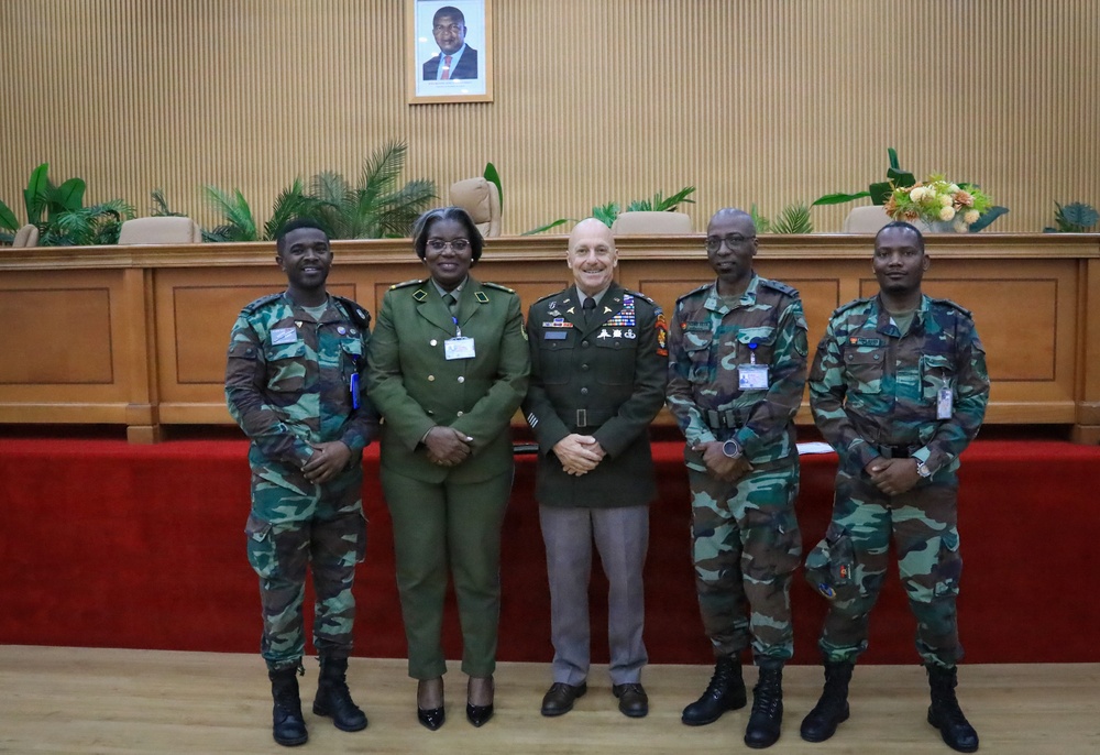 Five military personnel pose for a formal group photograph in what appears to be an official government or military conference room. The group includes individuals in different military uniforms, suggesting a multinational or joint military meeting. A framed portrait of an unidentified official hangs on the wall behind them.
