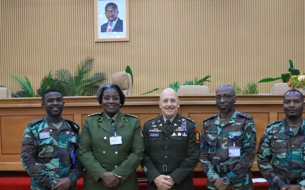 Five military personnel pose for a formal group photograph in what appears to be an official government or military conference room. The group includes individuals in different military uniforms, suggesting a multinational or joint military meeting. A framed portrait of an unidentified official hangs on the wall behind them.