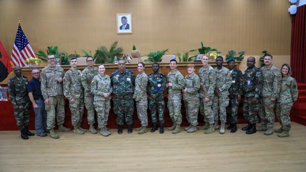 A group of approximately 16 military personnel and civilians pose for a formal group photograph in what appears to be an official government or military meeting hall. The group includes personnel in US Army MultiCam/OCP uniforms alongside individuals in different camouflage patterns, suggesting a multinational or bilateral military engagement. An American flag and what appears to be another nation's flag are visible in the background, along with a framed portrait on the wall.