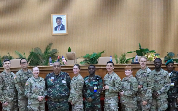 A group of approximately 16 military personnel and civilians pose for a formal group photograph in what appears to be an official government or military meeting hall. The group includes personnel in US Army MultiCam/OCP uniforms alongside individuals in different camouflage patterns, suggesting a multinational or bilateral military engagement. An American flag and what appears to be another nation's flag are visible in the background, along with a framed portrait on the wall.