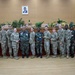 A group of approximately 16 military personnel and civilians pose for a formal group photograph in what appears to be an official government or military meeting hall. The group includes personnel in US Army MultiCam/OCP uniforms alongside individuals in different camouflage patterns, suggesting a multinational or bilateral military engagement. An American flag and what appears to be another nation's flag are visible in the background, along with a framed portrait on the wall.
