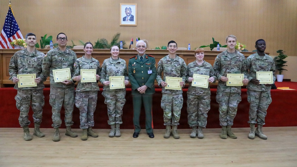 A group of eight military personnel pose for a formal photograph in what appears to be an official ceremony room. Seven individuals wearing US Army MultiCam/OCP camouflage uniforms are holding certificates, flanking a central figure in a distinctive green dress uniform with medals and decorations. The setting suggests a certificate presentation or graduation ceremony, likely in an official government or military facility.