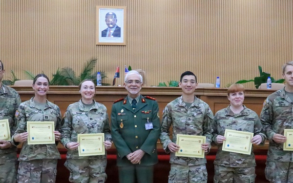 A group of eight military personnel pose for a formal photograph in what appears to be an official ceremony room. Seven individuals wearing US Army MultiCam/OCP camouflage uniforms are holding certificates, flanking a central figure in a distinctive green dress uniform with medals and decorations. The setting suggests a certificate presentation or graduation ceremony, likely in an official government or military facility.