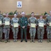 A group of eight military personnel pose for a formal photograph in what appears to be an official ceremony room. Seven individuals wearing US Army MultiCam/OCP camouflage uniforms are holding certificates, flanking a central figure in a distinctive green dress uniform with medals and decorations. The setting suggests a certificate presentation or graduation ceremony, likely in an official government or military facility.
