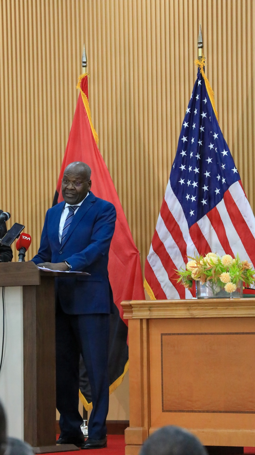 A man in a blue suit stands at a wooden podium delivering a speech or press conference. Behind him are two flags: the flag of Angola (red and black with a yellow emblem) and the flag of the United States of America. The setting appears to be a formal diplomatic or governmental venue, with a second podium visible to the right adorned with flowers.