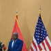 A man in a blue suit stands at a wooden podium delivering a speech or press conference. Behind him are two flags: the flag of Angola (red and black with a yellow emblem) and the flag of the United States of America. The setting appears to be a formal diplomatic or governmental venue, with a second podium visible to the right adorned with flowers.