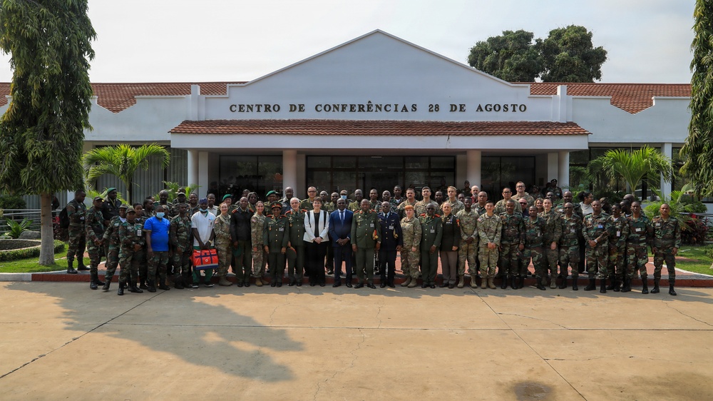 A large group of military personnel and civilians pose for a formal group photograph in front of the 'Centro de Conferências 28 de Agosto' (August 28 Conference Center). The group includes personnel in various military uniforms from what appear to be multiple nations, along with a few civilians in formal attire. The setting suggests a multinational military conference or cooperative training event.