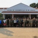 A large group of military personnel and civilians pose for a formal group photograph in front of the 'Centro de Conferências 28 de Agosto' (August 28 Conference Center). The group includes personnel in various military uniforms from what appear to be multiple nations, along with a few civilians in formal attire. The setting suggests a multinational military conference or cooperative training event.