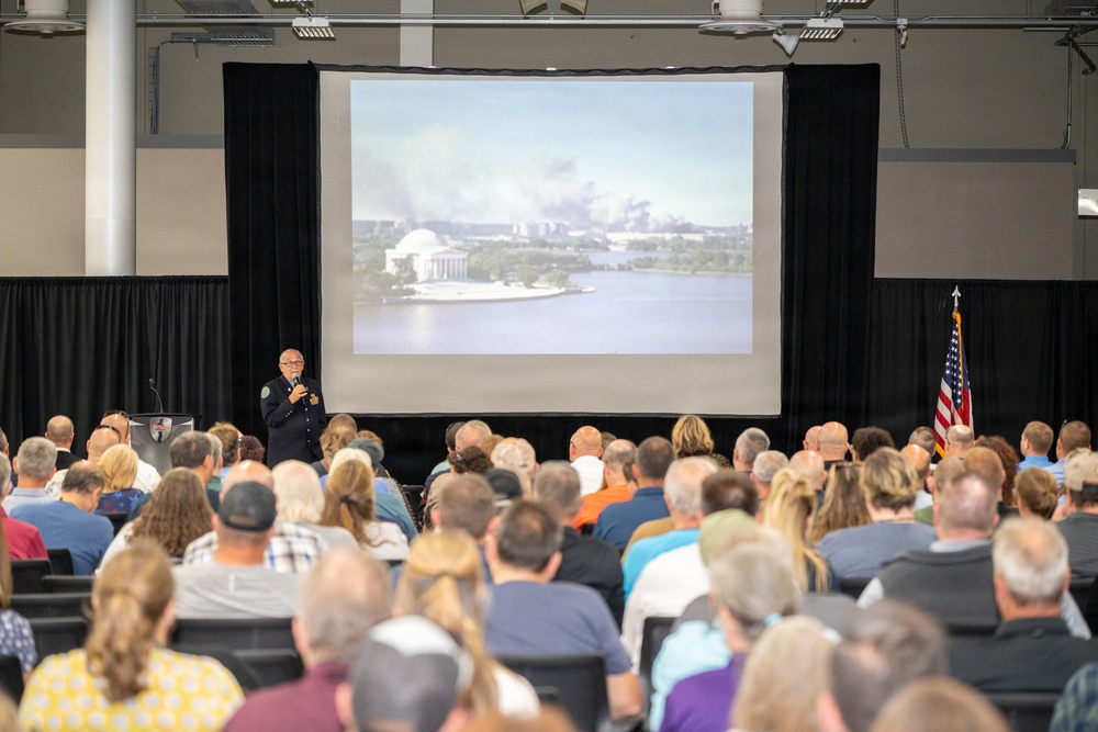 A uniformed official, likely a law enforcement or fire service officer, is presenting to a large seated audience in a convention-style hall. The projected image on the large screen behind the speaker shows the Jefferson Memorial in Washington D.C. with visible smoke rising in the background, suggesting the presentation may be related to the September 11, 2001 attacks or another significant emergency event. An American flag is displayed to the right of the screen.