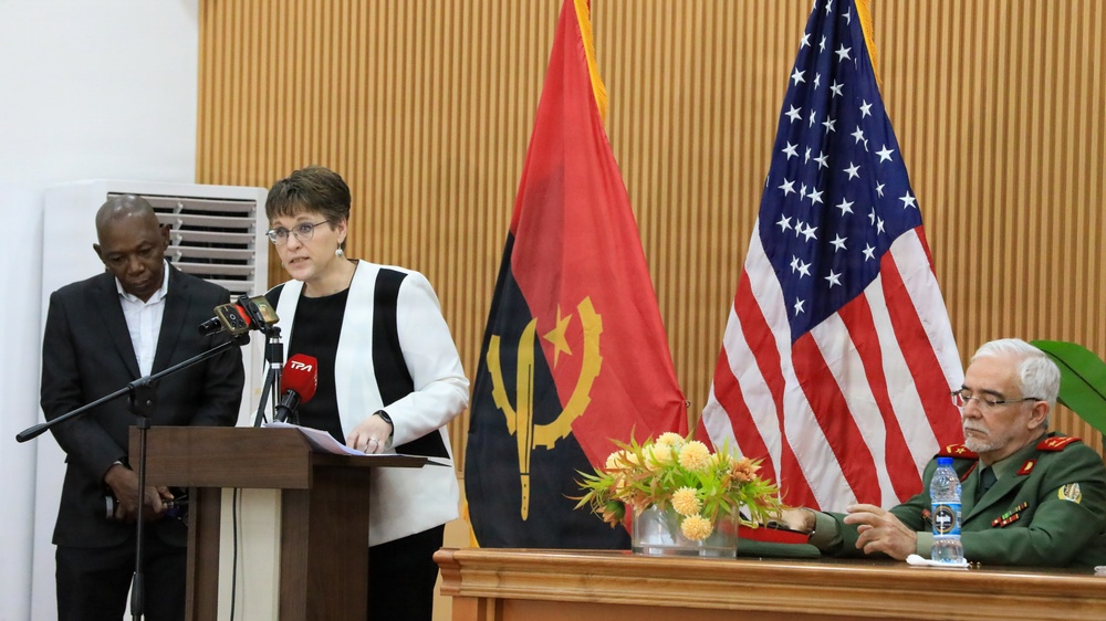 A formal diplomatic or official event featuring a woman speaking at a podium with microphones, flanked by the flags of Angola and the United States. A military officer in green uniform sits at a table to the right, while another individual stands to the left. The setting suggests a bilateral meeting, press conference, or official ceremony between Angola and the United States.