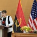 A formal diplomatic or official event featuring a woman speaking at a podium with microphones, flanked by the flags of Angola and the United States. A military officer in green uniform sits at a table to the right, while another individual stands to the left. The setting suggests a bilateral meeting, press conference, or official ceremony between Angola and the United States.