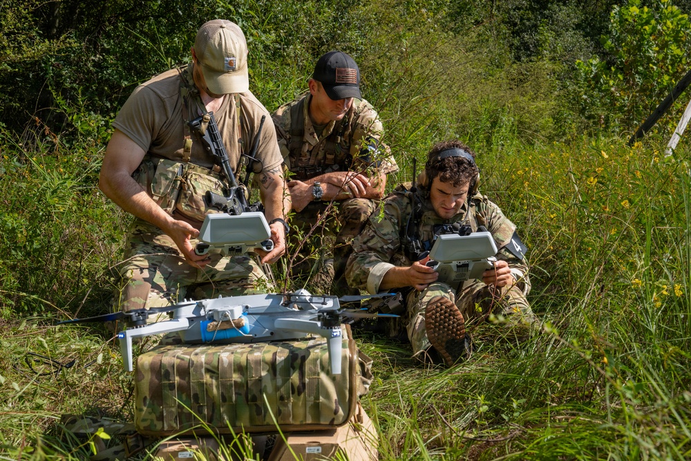 Three individuals in military camouflage uniforms are operating drone equipment in an outdoor, vegetated setting. Two of them are holding what appear to be drone controllers or tablets, while a quadcopter drone sits on a camouflage-patterned equipment bag in the foreground. The scene appears to depict a military or tactical drone operation or training exercise.