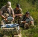 Three individuals in military camouflage uniforms are operating drone equipment in an outdoor, vegetated setting. Two of them are holding what appear to be drone controllers or tablets, while a quadcopter drone sits on a camouflage-patterned equipment bag in the foreground. The scene appears to depict a military or tactical drone operation or training exercise.