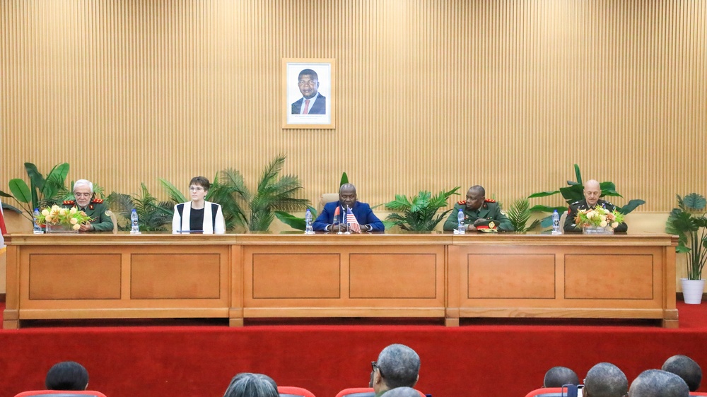 A formal diplomatic or military meeting is taking place in an official conference room. Five individuals are seated at a large wooden dais, including military officers in uniform and civilians, with small flags visible on the table. A framed portrait of an unidentified official hangs prominently on the wood-paneled wall behind the panel.