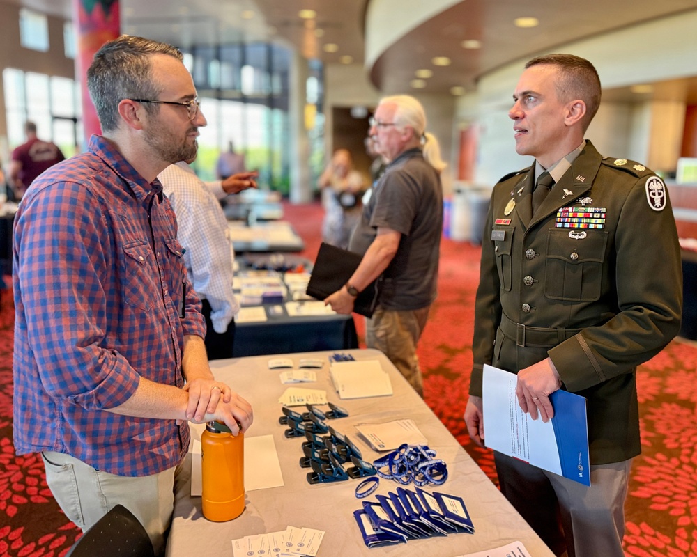 A U.S. Army officer in dress green uniform is engaged in conversation with a civilian man at what appears to be a career fair, expo, or outreach event held in a large conference or convention center. The officer is holding printed materials and wearing multiple medals and insignia. Several other attendees and exhibitor tables are visible in the background.