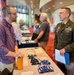 A U.S. Army officer in dress green uniform is engaged in conversation with a civilian man at what appears to be a career fair, expo, or outreach event held in a large conference or convention center. The officer is holding printed materials and wearing multiple medals and insignia. Several other attendees and exhibitor tables are visible in the background.