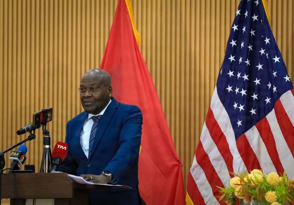 A man in a blue suit stands at a podium delivering a speech or press conference. The setting appears to be an official diplomatic or governmental event, as evidenced by the presence of both the United States flag and what appears to be the Angolan flag. Media microphones from outlets including 'TPA' and 'FAA' are visible at the podium.