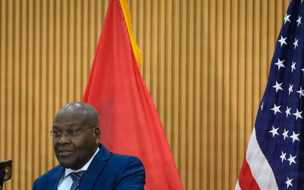 A man in a blue suit stands at a podium delivering a speech or press conference. The setting appears to be an official diplomatic or governmental event, as evidenced by the presence of both the United States flag and what appears to be the Angolan flag. Media microphones from outlets including 'TPA' and 'FAA' are visible at the podium.