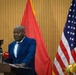 A man in a blue suit stands at a podium delivering a speech or press conference. The setting appears to be an official diplomatic or governmental event, as evidenced by the presence of both the United States flag and what appears to be the Angolan flag. Media microphones from outlets including 'TPA' and 'FAA' are visible at the podium.