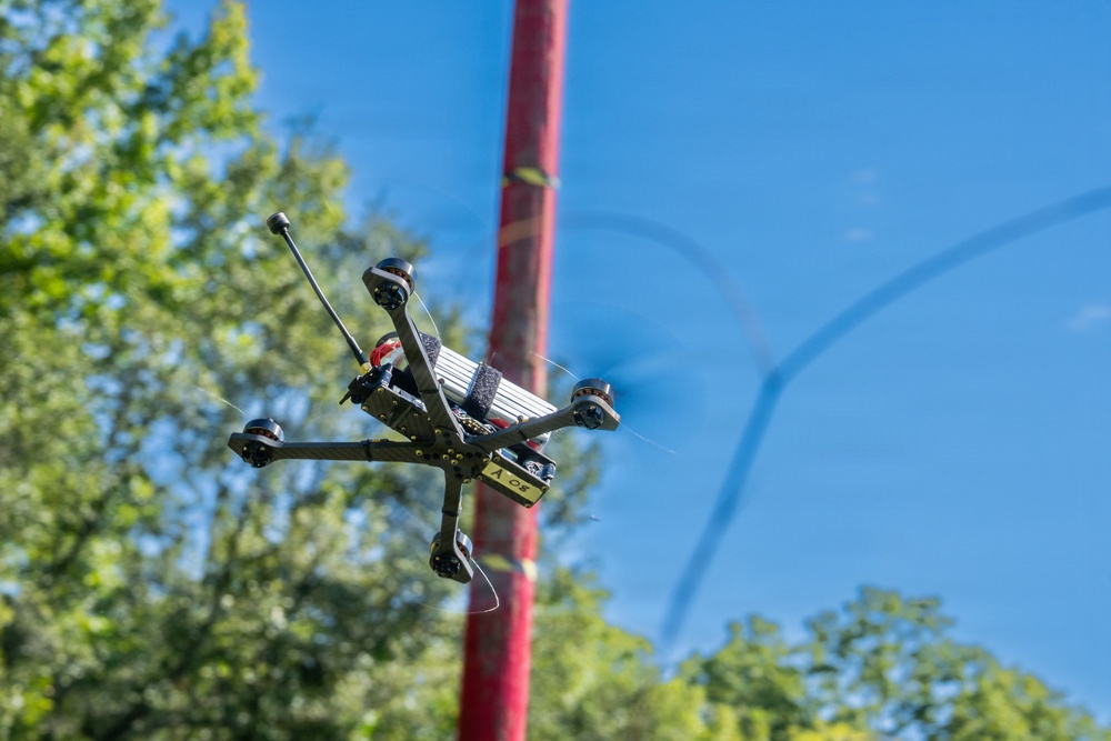 An FPV (First Person View) racing drone is captured mid-flight against a clear blue sky. The drone's propellers are visibly spinning, shown by motion blur, and it appears to be maneuvering near a red pole or post. Green trees are visible in the background, suggesting an outdoor recreational or racing environment.
