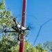 An FPV (First Person View) racing drone is captured mid-flight against a clear blue sky. The drone's propellers are visibly spinning, shown by motion blur, and it appears to be maneuvering near a red pole or post. Green trees are visible in the background, suggesting an outdoor recreational or racing environment.