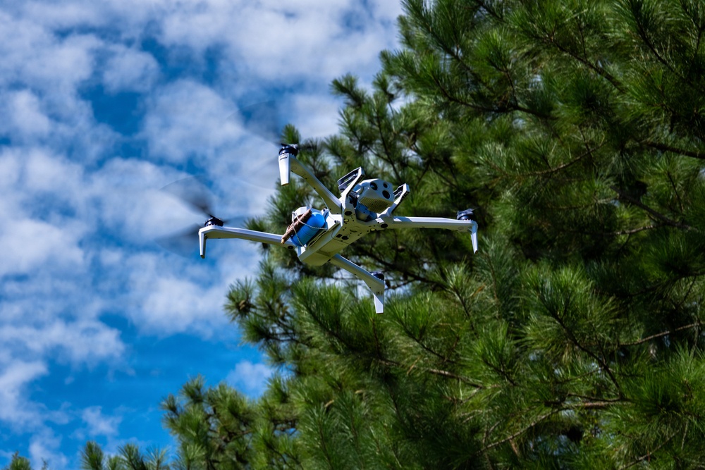 A multi-rotor drone is captured mid-flight against a partly cloudy blue sky, with pine trees visible in the background and foreground. The drone appears to be a custom or specialized hexacopter-style UAV with a white frame, featuring a blue cylindrical payload or component mounted at its center. The image is taken from a low angle looking upward, providing a clear view of the drone's underside and rotor configuration.